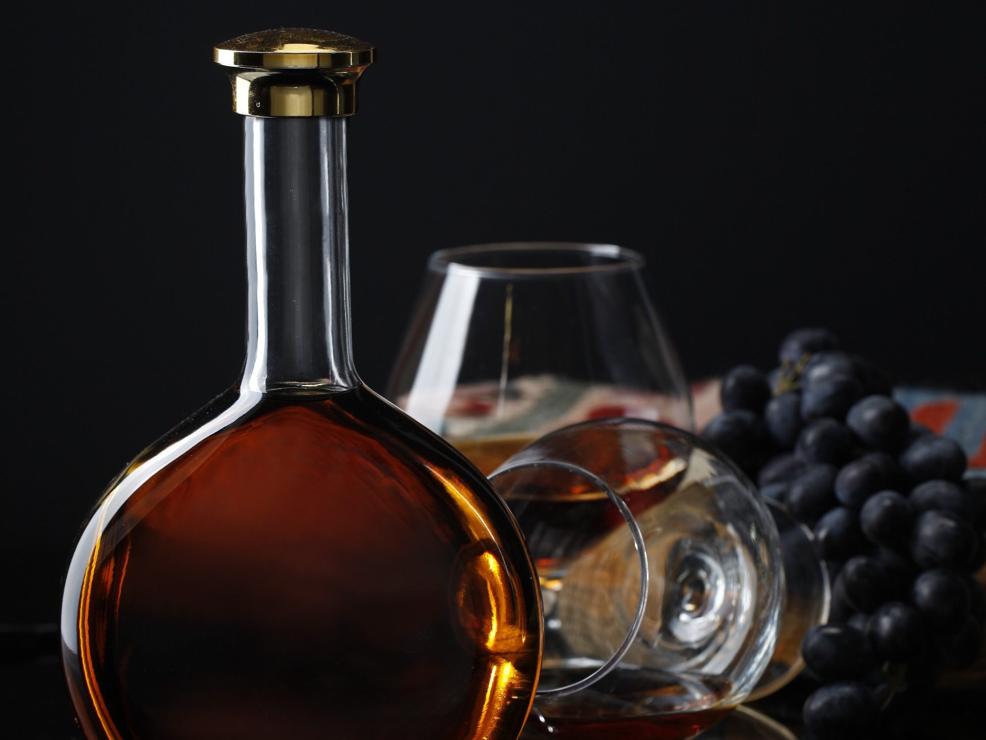 Glazen fles met amberkleurige drank, een whiskyglas en druiven op donkere achtergrond.