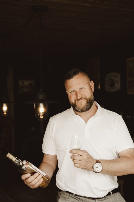 Man met een baard in een lichte blouse houdt een fles en een glas in een donkere ruimte.